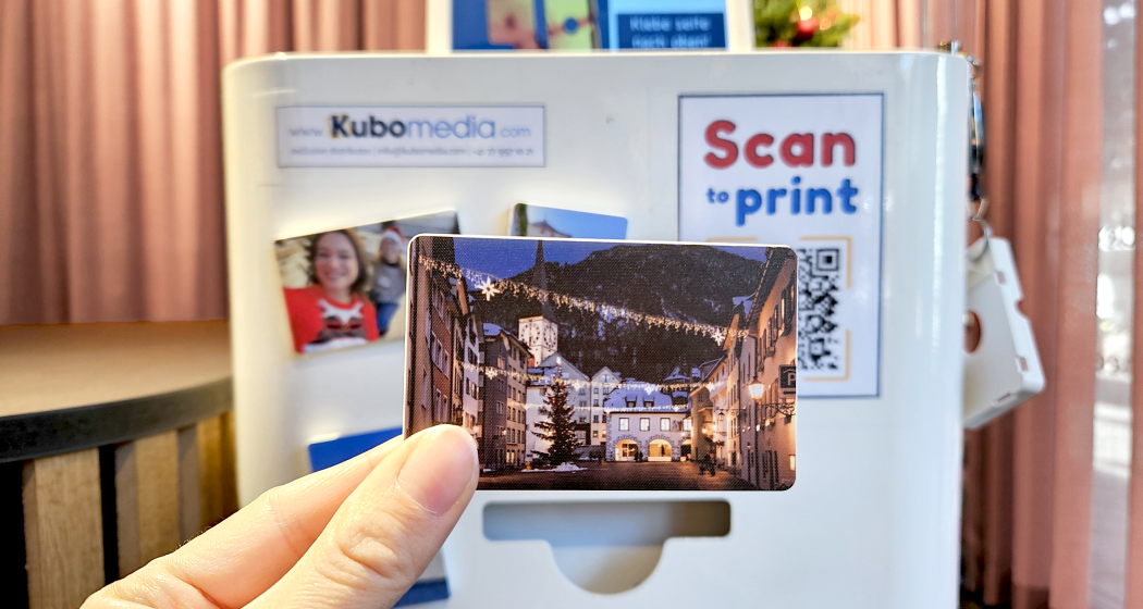 Fotomagnetdrucker (© Chur Tourismus) Eine Hand hält ein Magnet mit einem Foto von der weihnachtlich beleuchteten Altstadt, im Hintergrund ist der Fotomagnetdrucker