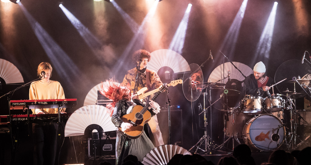 Arcas Rock (© Arcas Rock) Eine Band aus 4 Personen tritt auf der Bühne auf. Helle Scheinwerfer strahlen von oben, und weiße Fächer schmücken den Hintergrund.