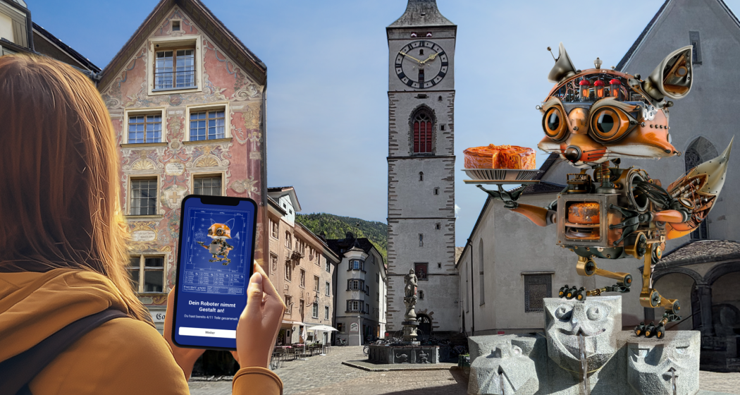 Foxtrail GO Chur (©Foxtrail) Eine Person hält ein Smartphone in der Hand, auf dem ein Roboterfuchs zu sehen ist, passend zu einer grossen Roboterfuchs-Skulptur, in Chur mit historischen Gebäuden und einem Uhrturm.