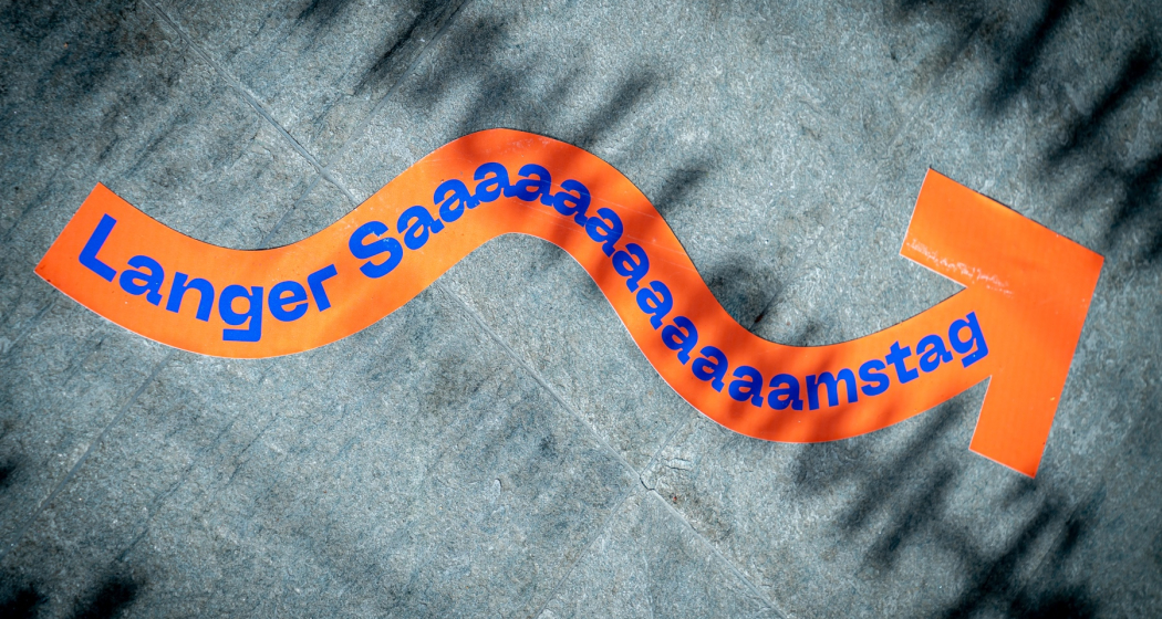 Langer Samstag 2025 (© Yanik Bürkli) Ein orangefarbener, geschwungener Aufkleber auf dem Boden zeigt den Text „Langer Saaamaaaamstag“ in blauer Schrift und weist mit einem Pfeil die Richtung. Das Licht wirft Schatten auf den Steinboden und betont die verspielte Gestaltung.