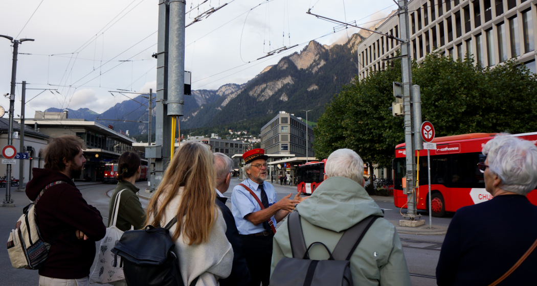 Auf Durchzug (© Chur Tourismus) Eine Gruppe von Menschen hören einem Stadtführer im historischen Bahnhofsoutfit aufmerksam zu. Im Hintergrund sind der Bahnhof Chur, rote Stadtbusse und die umliegende Berglandschaft zu sehen.