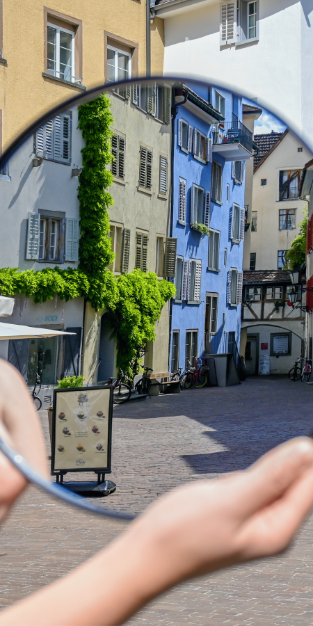 Im Hintergrund ist die Altstadt zu sehen, im Vordergrund hält eine Person einen Spiegel, wo sich ebenfalls die Altstadt spiegelt