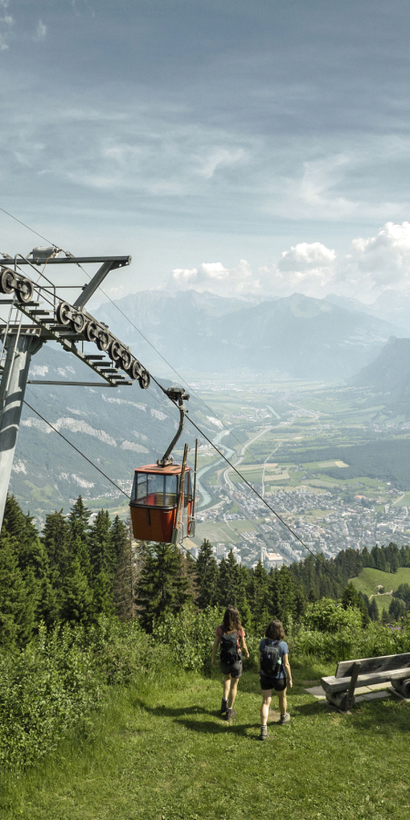 Two people walk toward a cable car in a scenic mountain landscape. A Swiss flag flutters nearby, with lush greenery and a town below