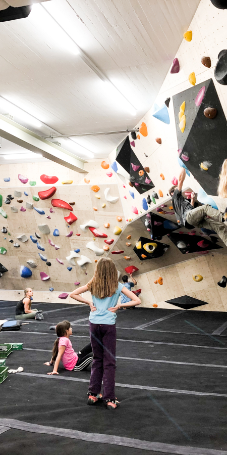Boulder Quadrel (© Quadrel) Children bouldering on colorful climbing walls in an indoor gym. Others sit and watch on padded floors, creating a playful and adventurous atmosphere