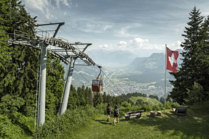 Two people walk toward a cable car in a scenic mountain landscape. A Swiss flag flutters nearby, with lush greenery and a town below