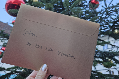 Eine Hand hält einen goldenen Briefumschlag mit der Aufschrift ‚juhui, du hast mich gefunden.‘ vor einem geschmückten Weihnachtsbaum mit roten und goldenen Kugeln.