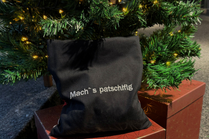 Schwarzes Wärmekissen mit weissem Schriftzug ‚Mach’s patschifig‘, platziert auf einem roten Geschenkpaket unter einem beleuchteten Weihnachtsbaum.