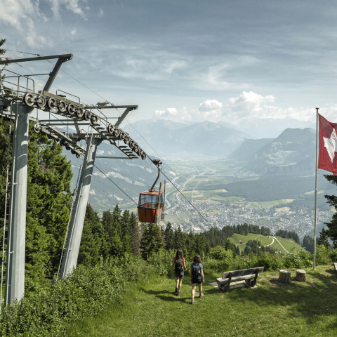 Two people walk toward a cable car in a scenic mountain landscape. A Swiss flag flutters nearby, with lush greenery and a town below