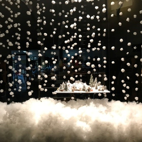 Winterliche Schaufensterdekoration mit einer kleinen verschneiten Landschaft aus Tannenbäumen und Häusern, umgeben von schwebenden Watte-Schneeflocken und einer dichten Schneeschicht am Boden.