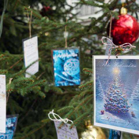Weihnachtskarten und Wunschzettel hängen an den Zweigen eines festlich geschmückten Tannenbaums im Freien; im Hintergrund sind rote Kugeln und unscharfe Stadtfassaden zu erkennen.
