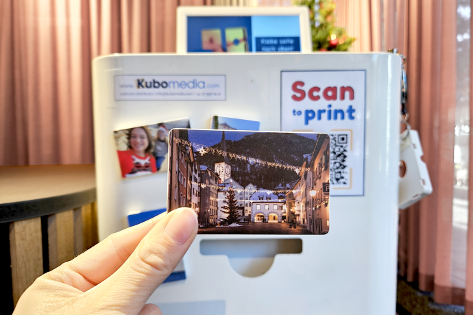 Fotomagnetdrucker (© Chur Tourismus) Eine Hand hält ein Magnet mit einem Foto von der weihnachtlich beleuchteten Altstadt, im Hintergrund ist der Fotomagnetdrucker
