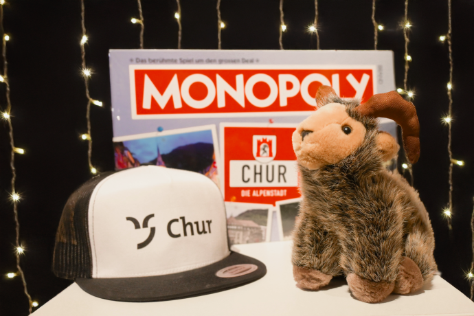 Monopoly, Mütze mit Chur Logo und ein Steinbock als Plüschtier vor einem leuchtenden Hintergrund