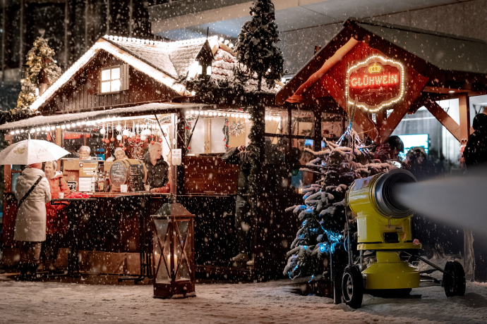 2 Stände auf dem Weihnachtsmarkt, bei den Ständen stehen Menschen, eine Schneekanone produziert Schnee