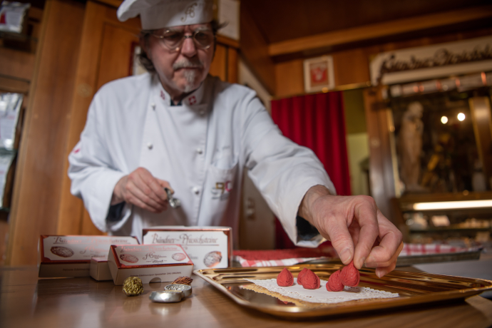 Ein Koch in weißer Uniform und Kochmütze platziert rote Pfirsichsteine auf einem goldenen Tablett in einer rustikalen Umgebung.
