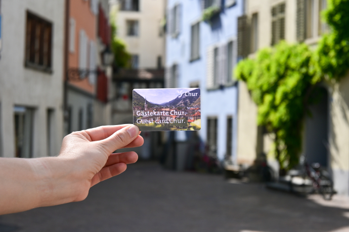Gästekarte Chur Im Hintergrund sind Stadtmauern zu sehen, im Vordergrund hält eine Person die Gästekarte von Chur in den Händen.