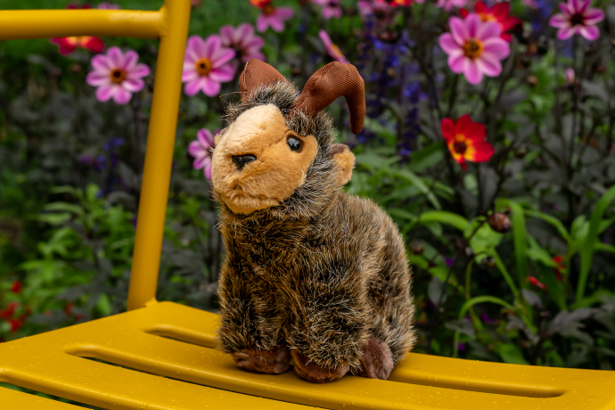 Ein Plüschtier in Form eines Steinbocks sitzt auf einem gelben Stuhl. Im Hintergrund blühen pinke und rote Blumen, die der Szene einen farbenfrohen, sommerlichen Rahmen geben.