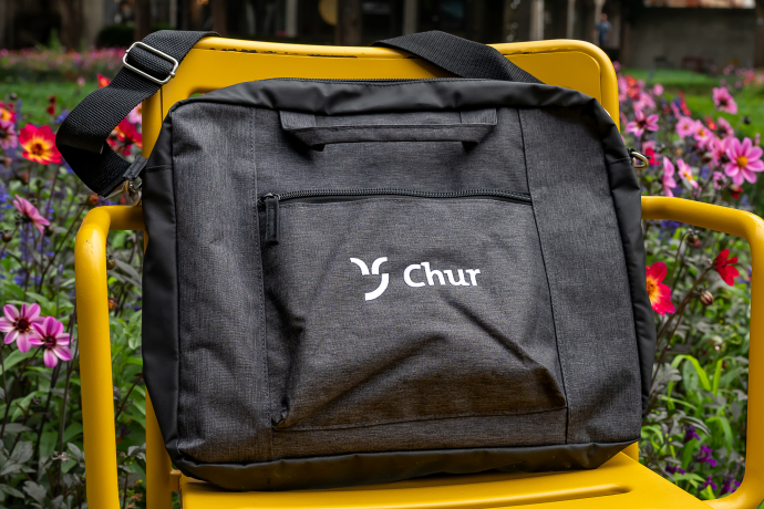 Eine schwarze Umhängetasche mit dem "Chur Tourismus"-Logo liegt auf einem gelben Stuhl. Im Hintergrund sorgen bunte Blumen für einen farbenfrohen Kontrast zur schlichten Tasche.