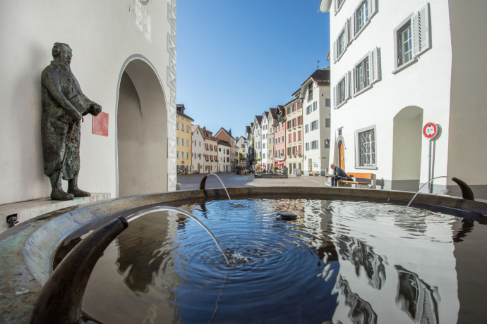 Historic European street with colorful buildings under a clear blue sky. Foreground features a rippling fountain near a bronze statue, creating a serene atmosphere