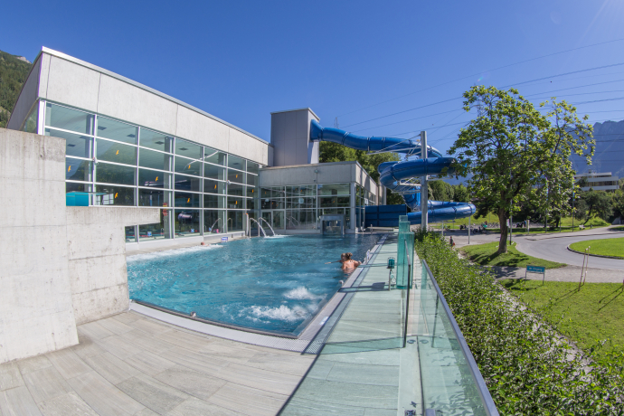 Modern aquatic center with an outdoor pool, glass walls, and a blue waterslide. A few people relax in the sun, surrounded by greenery