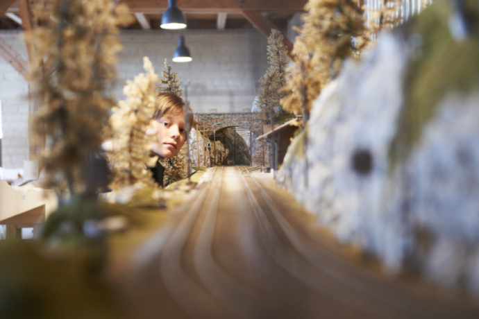 A child intently observes a detailed miniature train model with trees and a tunnel in a warmly lit room, evoking curiosity and wonder