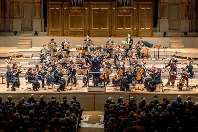 An orchestra performs on a grand stage with musicians playing strings, brass, and percussion. The conductor leads passionately; audience attentively watches