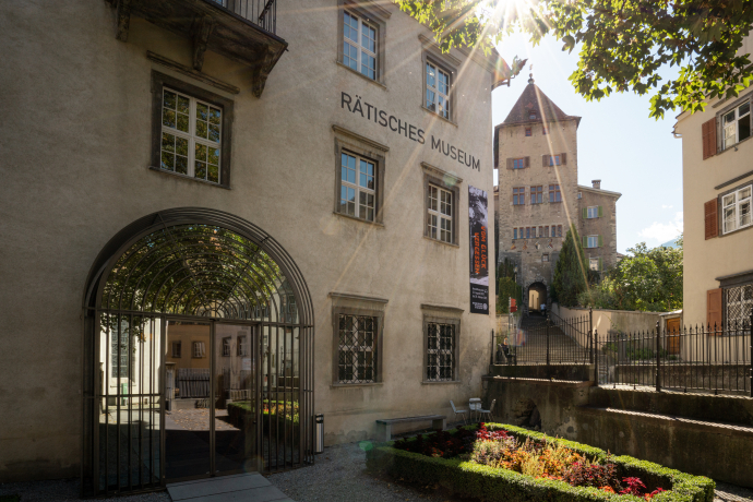 Sunlit facade of Rätisches Museum with arched entrance, surrounded by manicured gardens. A historic tower rises in the background under a clear sky