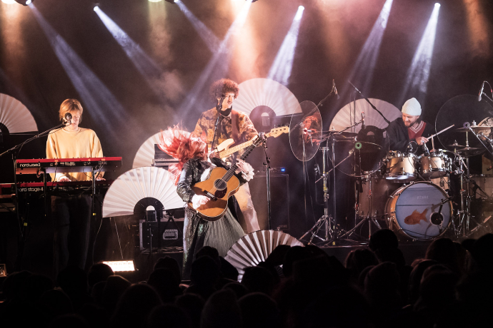 Eine Band aus 4 Personen tritt auf der Bühne auf. Helle Scheinwerfer strahlen von oben, und weiße Fächer schmücken den Hintergrund.
