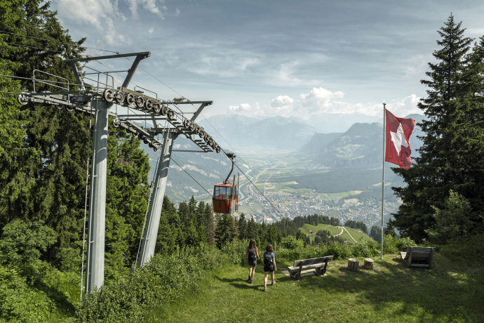 Two people walk toward a cable car in a scenic mountain landscape. A Swiss flag flutters nearby, with lush greenery and a town below