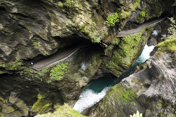 A dramatic overhead view of a narrow mountain gorge with a winding walkway. Lush greenery clings to rocky walls above turquoise rapids below
