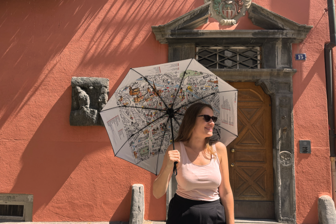 Churer Regenschirm (©Chur Tourismus)  Eine Frau steht vor einer historischen Hausfassade mit roter Wand und altem Holztor. Sie trägt Sonnenbrille, helles Top und dunkle Hose, während sie einen aufgespannten Regenschirm mit Stadtplan-Motiv in der Hand hält und lächelnd zur Seite blickt.