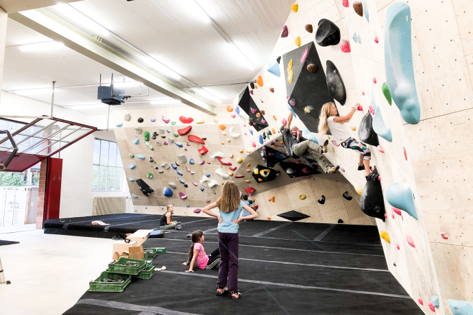 Boulder Quadrel (© Quadrel) Children bouldering on colorful climbing walls in an indoor gym. Others sit and watch on padded floors, creating a playful and adventurous atmosphere