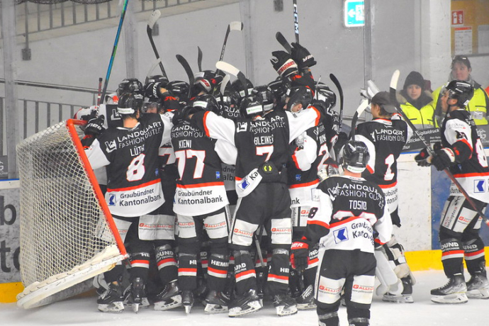 A hockey team huddles joyfully in front of a goal, raising their sticks. They wear black and white uniforms with red accents. The mood is celebratory