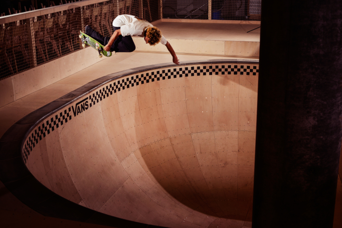 A skateboarder in mid-air performs a stylish trick above a deep, empty skate bowl with a Vans logo