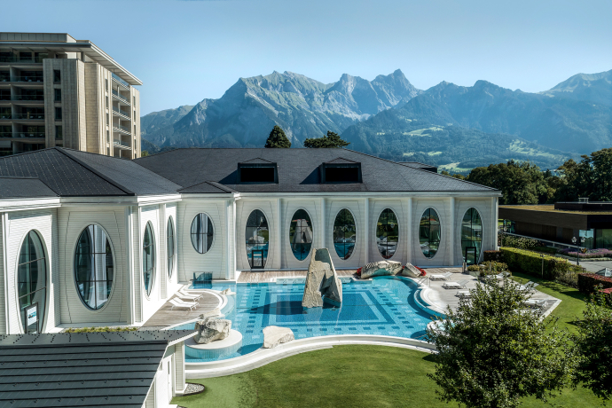 A luxurious spa building with large oval windows overlooks a scenic mountain range. The blue pool is surrounded by lounging areas