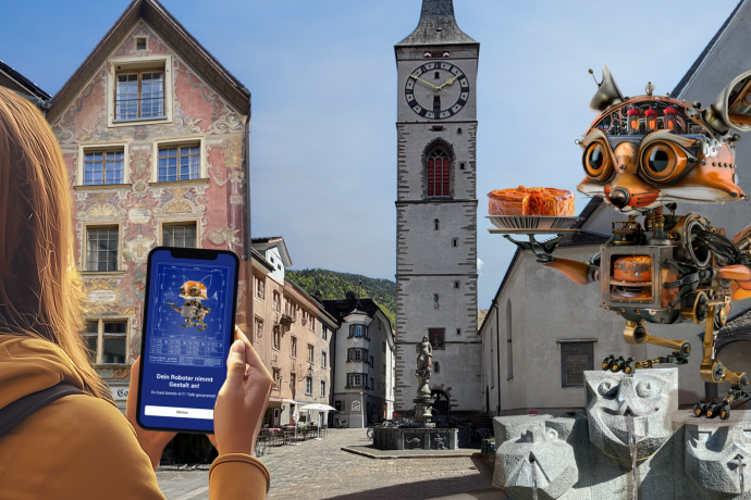 Eine Person hält ein Smartphone in der Hand, auf dem ein Roboterfuchs zu sehen ist, passend zu einer grossen Roboterfuchs-Skulptur, in Chur mit historischen Gebäuden und einem Uhrturm.