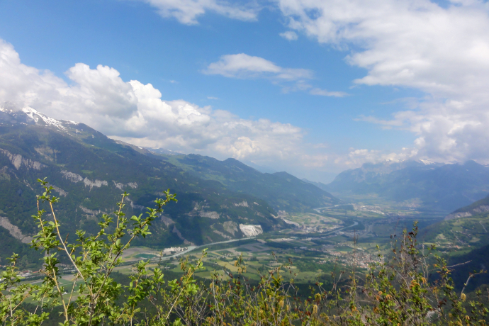 Ein weiter Blick über das Churer Rheintal mit seinen grünen Feldern und der sich schlängelnden Rheinlinie. Umgeben von hohen Bergen mit teils schneebedeckten Gipfeln breitet sich die Landschaft unter einem leicht bewölkten Sommerhimmel aus.