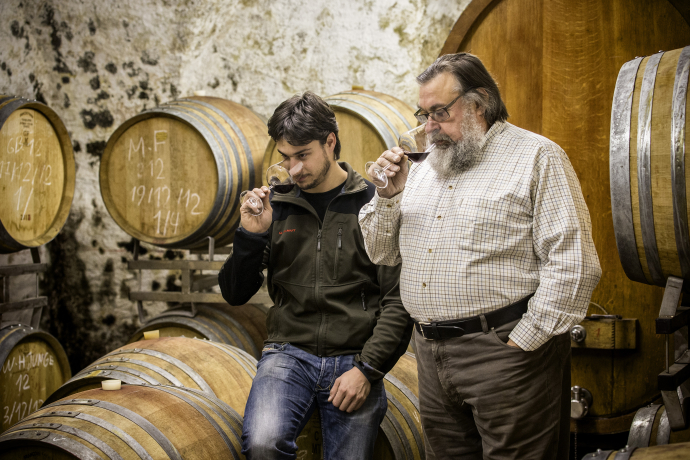 Zwei Männer stehen in einem Weinkeller vor grossen Holzfässern. Sie halten jeweils ein Glas Rotwein in der Hand und prüfen konzentriert den Duft. Die Szene vermittelt Handwerkstradition und die Kunst der Weinverkostung.