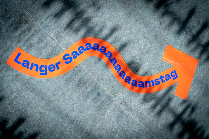 Langer Samstag 2025 (© Yanik Bürkli) Ein orangefarbener, geschwungener Aufkleber auf dem Boden zeigt den Text „Langer Saaamaaaamstag“ in blauer Schrift und weist mit einem Pfeil die Richtung. Das Licht wirft Schatten auf den Steinboden und betont die verspielte Gestaltung.