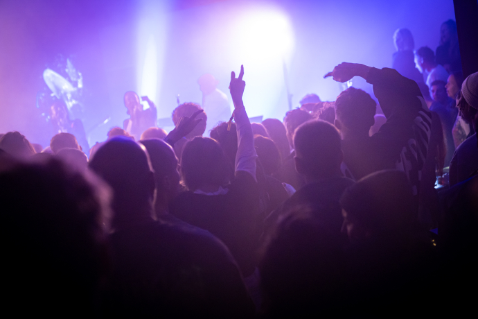 Langer Samstag, Werkstatt (© Yanik Bürkli)  Eine dichte Menschenmenge tanzt und feiert in stimmungsvoller Club-Atmosphäre. Bunte Lichter und Nebel tauchen die Szene in ein violett-blaues Licht, während auf der Bühne Musiker auftreten und das Publikum mitgerissen die Hände hebt.