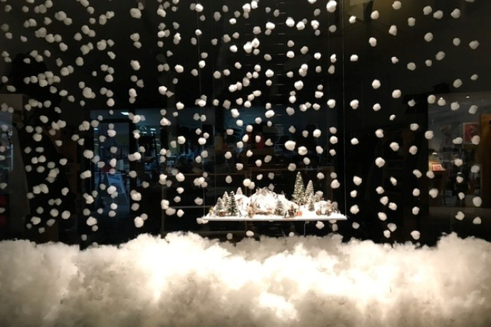 Winterliche Schaufensterdekoration mit einer kleinen verschneiten Landschaft aus Tannenbäumen und Häusern, umgeben von schwebenden Watte-Schneeflocken und einer dichten Schneeschicht am Boden.
