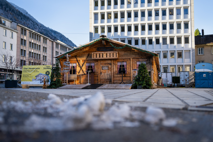 Eine gemütliche Alphütte steht auf dem Theaterplatz in Chur, umgeben von städtischen Gebäuden und mit Blick auf verschneite Berghänge. Vor der Hütte stehen Tische und kleine Tannenbäume, ein Schild kündigt Käsefondue und Raclette an
