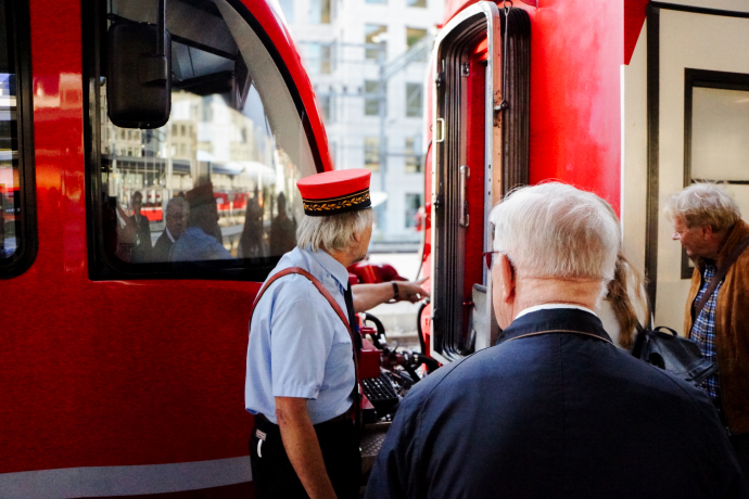 Auf Durchzug 2 (© Chur Tourismus) ChatGPT: Ein Stadtführer in historischer Uniform mit roter Mütze zeigt einer Gruppe von Besuchern Details an einem roten Zug der Rhätischen Bahn. Die Szene spielt am Bahnhof ab.