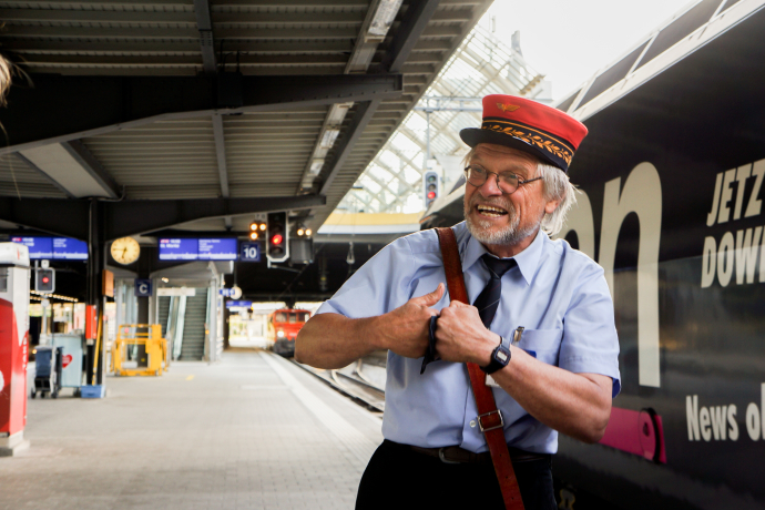 Auf Durchzug 3 (© Chur Tourismus) Ein Stadtführer in historischer Uniform mit roter Mütze steht lachend am Bahnsteig. Hinter ihm sind ein Zug, Bahnsteigschilder und die Bahnhofsuhr zu sehen.