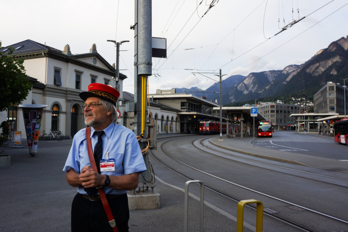 Auf Durchzug 5 (© Chur Tourismus) Ein Stadtführer in historischer Uniform mit roter Mütze steht vor dem Bahnhof Chur. Hinter ihm sind Busse und Züge sowie die umliegende Berglandschaft sichtbar, was der Szene einen authentischen alpinen Rahmen gibt.