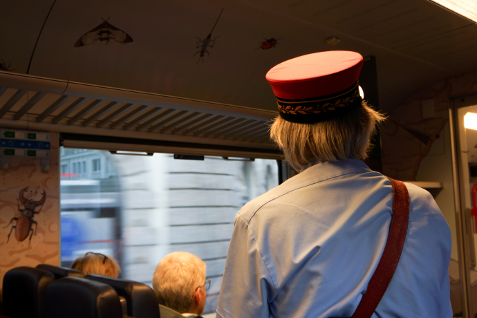 Auf Durchzug 3 (© Chur Tourismus) Ein Stadtführer in historischer Uniform mit roter Mütze steht im Inneren der Arosabahn. Er blickt aus dem Zugfenster, während vor ihm Fahrgäste sitzen. An den Wänden sind Tierillustrationen zu sehen, die den Wagen schmücken.