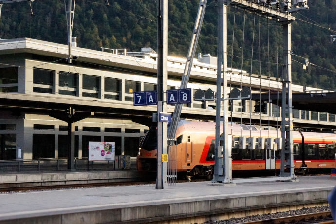 Bahnhof (© Chur Tourismus) Ein moderner Zug steht am Bahnhof Chur. Im Vordergrund sind die Gleise sowie die Beschilderung der Bahnsteige 7 und 8 zu sehen, im Hintergrund das Bahnhofsgebäude und bewaldete Hänge.