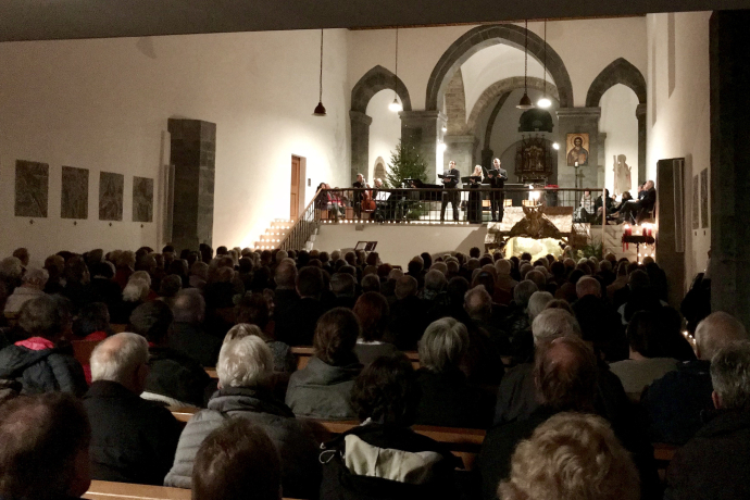 Weihnachtskonzert in der Kirche St. Luzi in Chur: Ein Ensemble aus Sängerinnen, Sängern und Musikerinnen tritt vor vollbesetztem Publikum auf. Im Hintergrund sind Weihnachtsbaum, Krippe und Kirchenarchitektur stimmungsvoll beleuchtet.