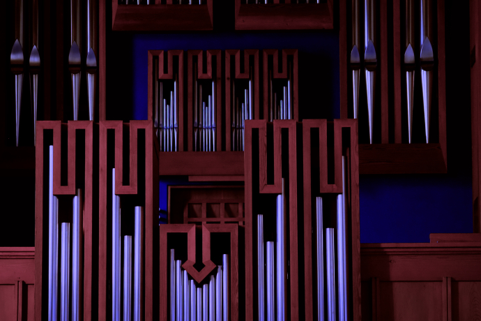 Detailansicht einer Kirchenorgel mit roten Holzrahmen und silbrig glänzenden Pfeifen vor blauem Hintergrund, kunstvoll beleuchtet.