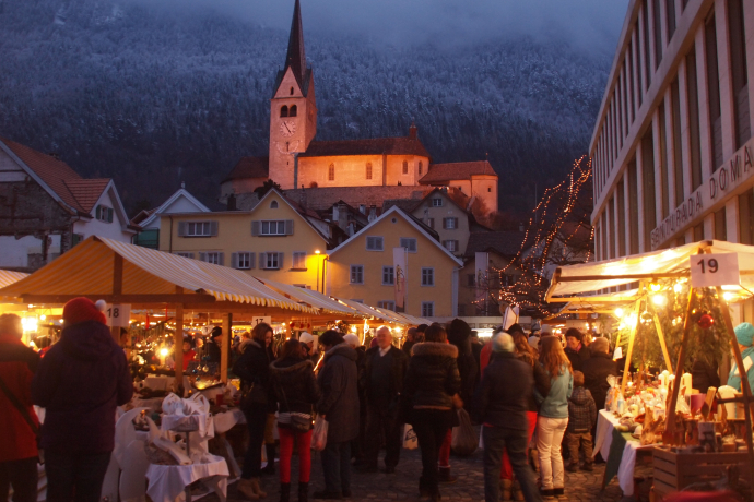 Abendlicher Weihnachtsmarkt in Domat/Ems mit festlich geschmückten Ständen, vielen Besuchern und der beleuchteten Kirche im Hintergrund vor verschneiten Bergen.