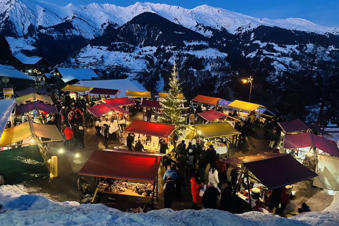 Stimmungsvoller Weihnachtsmarkt in Tschiertschen mit bunt beleuchteten Ständen, Besuchern und einem geschmückten Weihnachtsbaum, umgeben von verschneiten Bergen und winterlicher Abendstimmung.